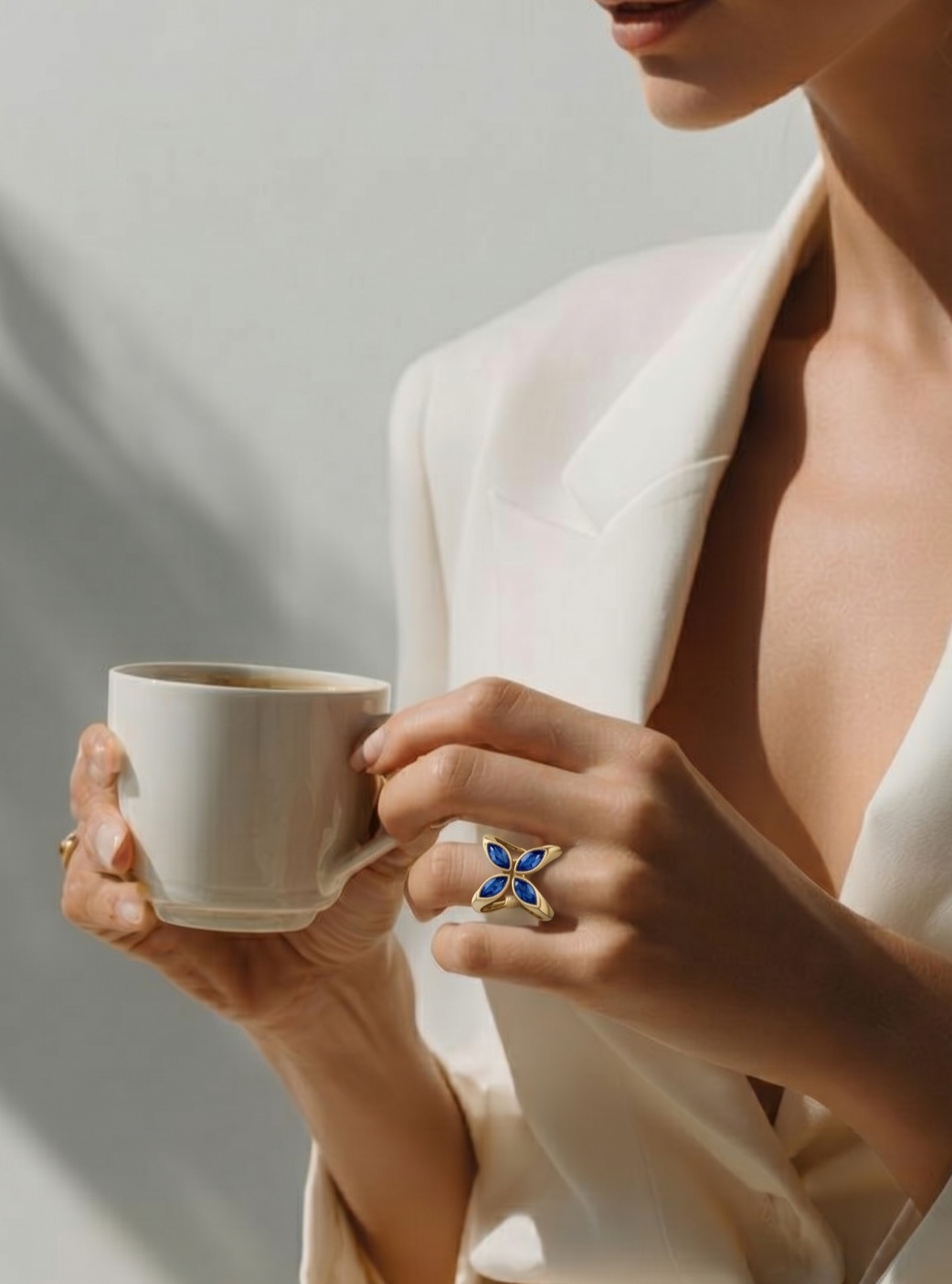 A hand wearing the Quatrefoil Ring — four marquise natural blue sapphires in champagne gold — holding a white coffee cup in soft morning light.